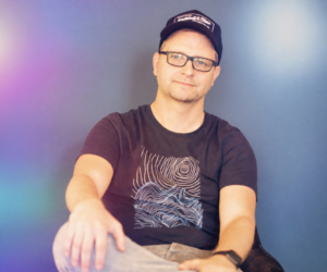 Mann mit Brille und Kapuzenmütze sitzt entspannt vor einem farbigen Hintergrund.